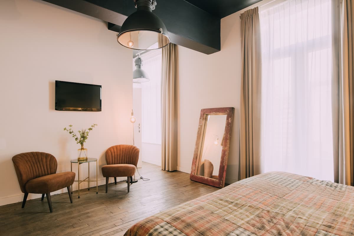 A hotel room with a bed, two armchairs, a small table with a vase, a TV, a mirror, and a large window with curtains.