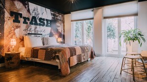 A bedroom with a large bed, a nightstand with a lamp, a wall mural, and a large window with blinds. - Trash Deluxe (Maastricht)