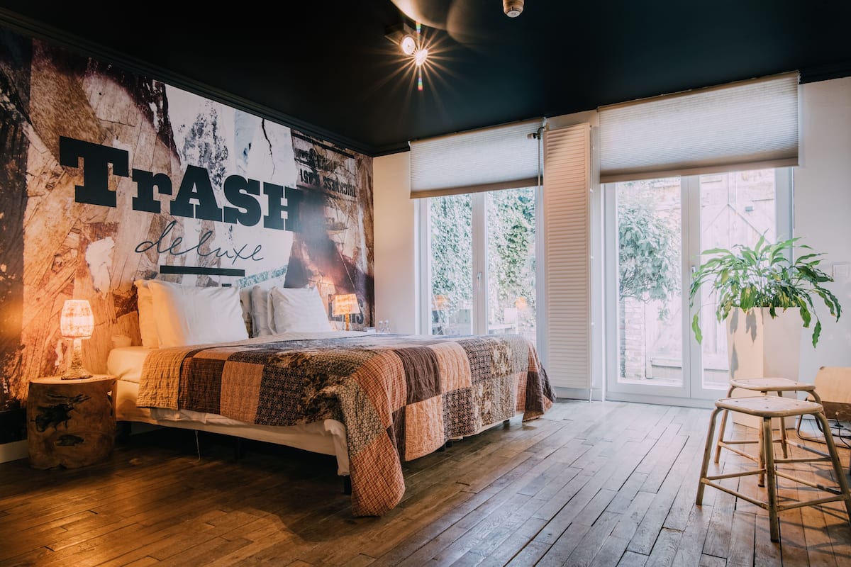 A bedroom with a large bed, a nightstand with a lamp, a wall mural, and a large window with blinds.