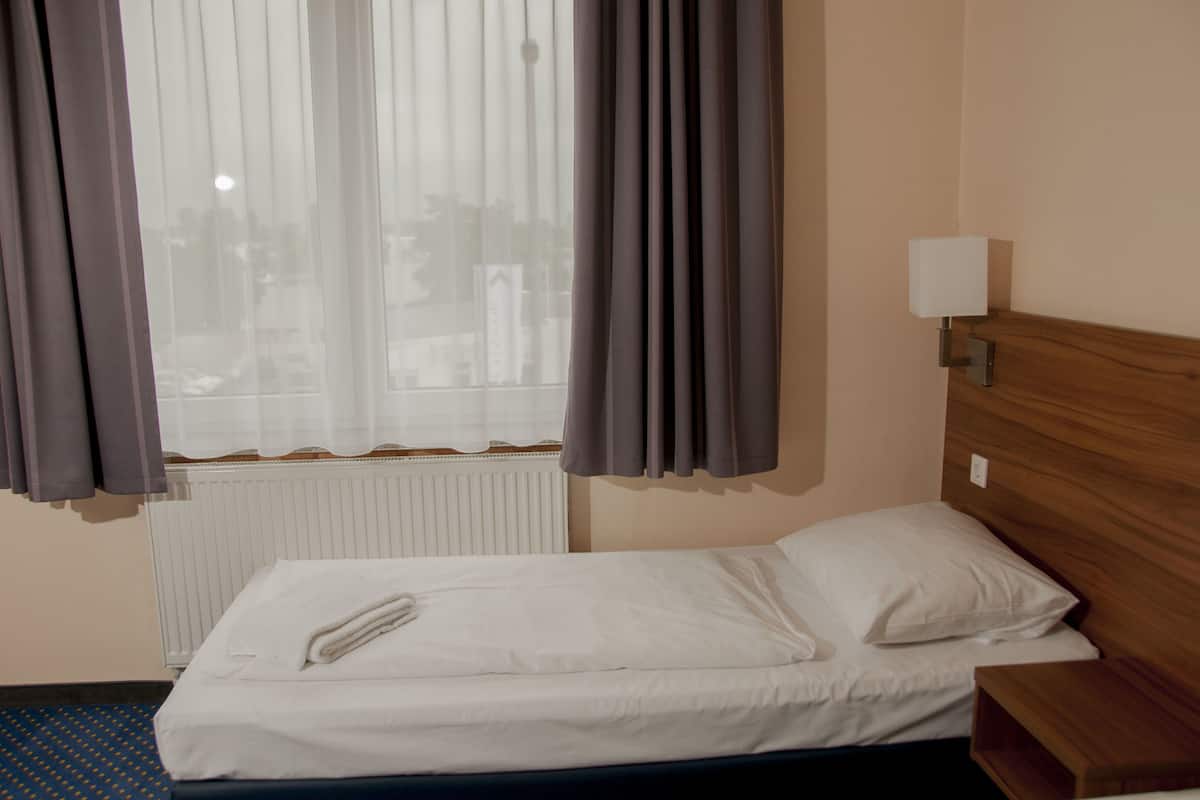 Une chambre d’hôtel avec un lit simple, une tête de lit en bois, une table de chevet, une lampe et une fenêtre avec des rideaux.
