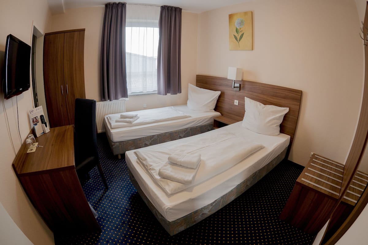 Une chambre d’hôtel avec deux lits simples, un bureau avec une chaise, un téléviseur fixé au mur et une fenêtre avec des rideaux.