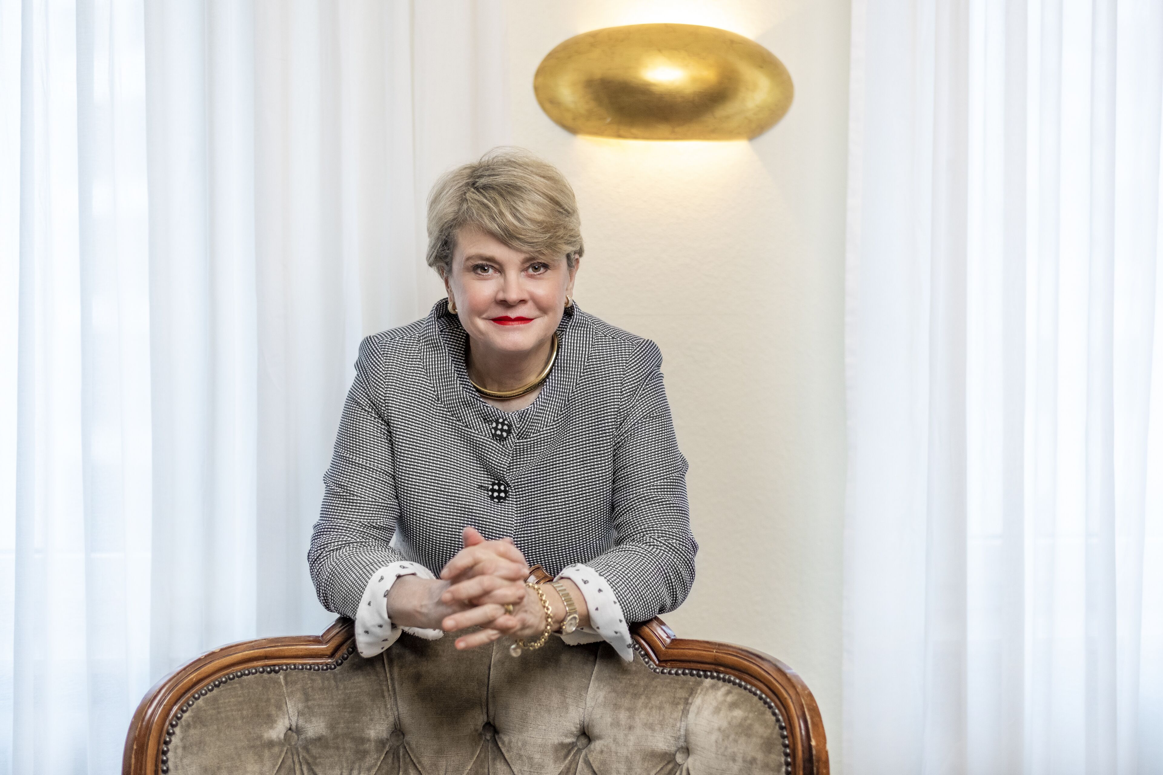 A person seated on a tufted chair with a patterned jacket, in a room with white curtains and a wall-mounted light fixture.