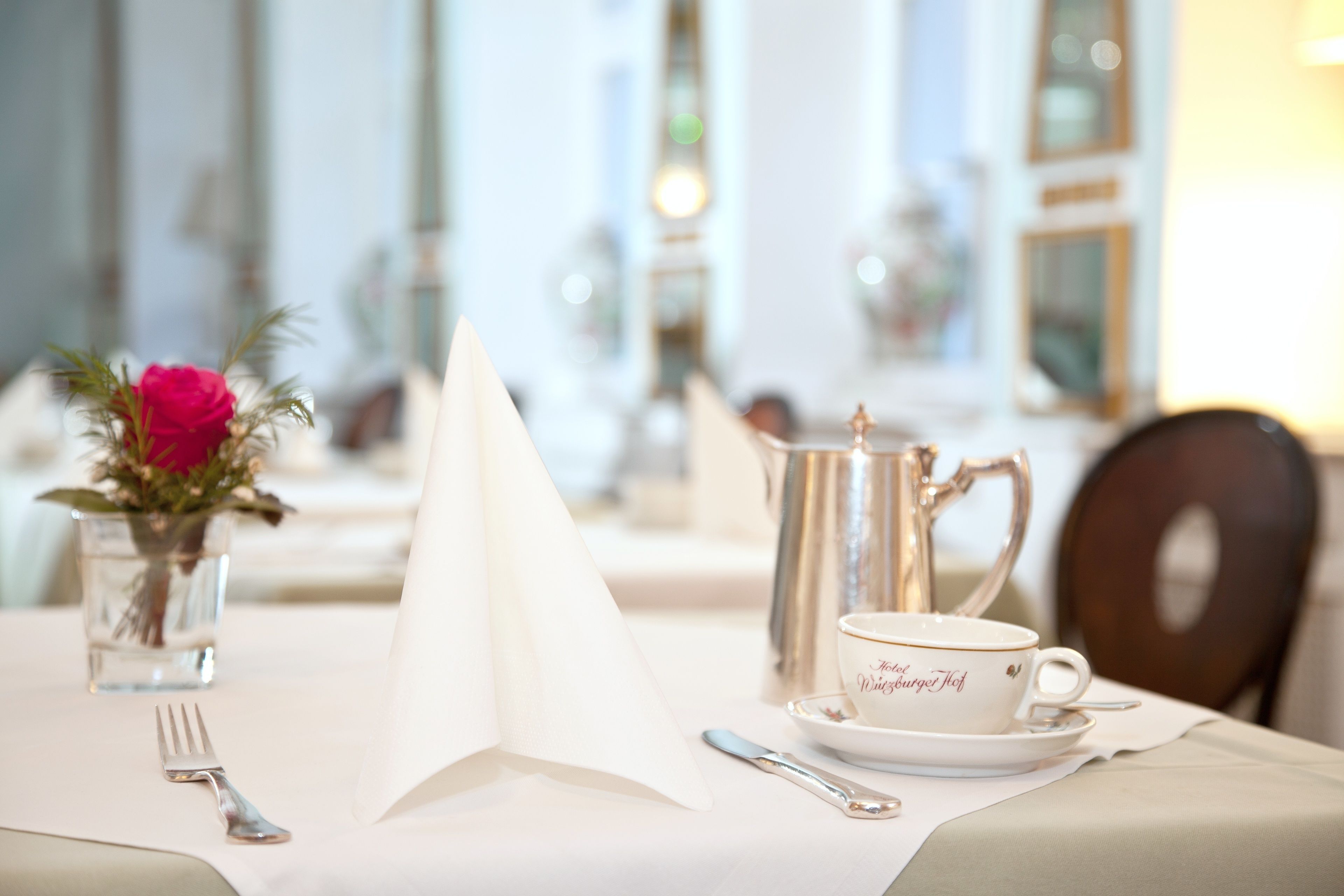 A dining table set with a white tablecloth, a silver pitcher, a white cup with gold lettering, a fork, and a knife.