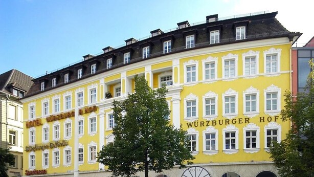 A yellow building with the sign "WURZBURGER HOF" indicating a hotel. There are people walking around, and a tree in the center. - Hotel Würzburger Hof (Würzburg)
