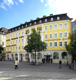 A yellow building with the sign "WURZBURGER HOF" indicating a hotel. There are people walking around, and a tree in the center. - Hotel Würzburger Hof (Würzburg)