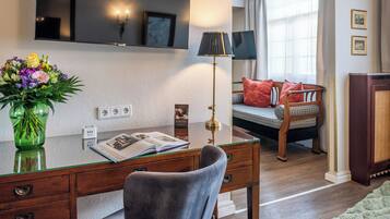 A hotel room with a wooden desk, a chair, a bed with red pillows, a lamp, a TV, and a vase with flowers.