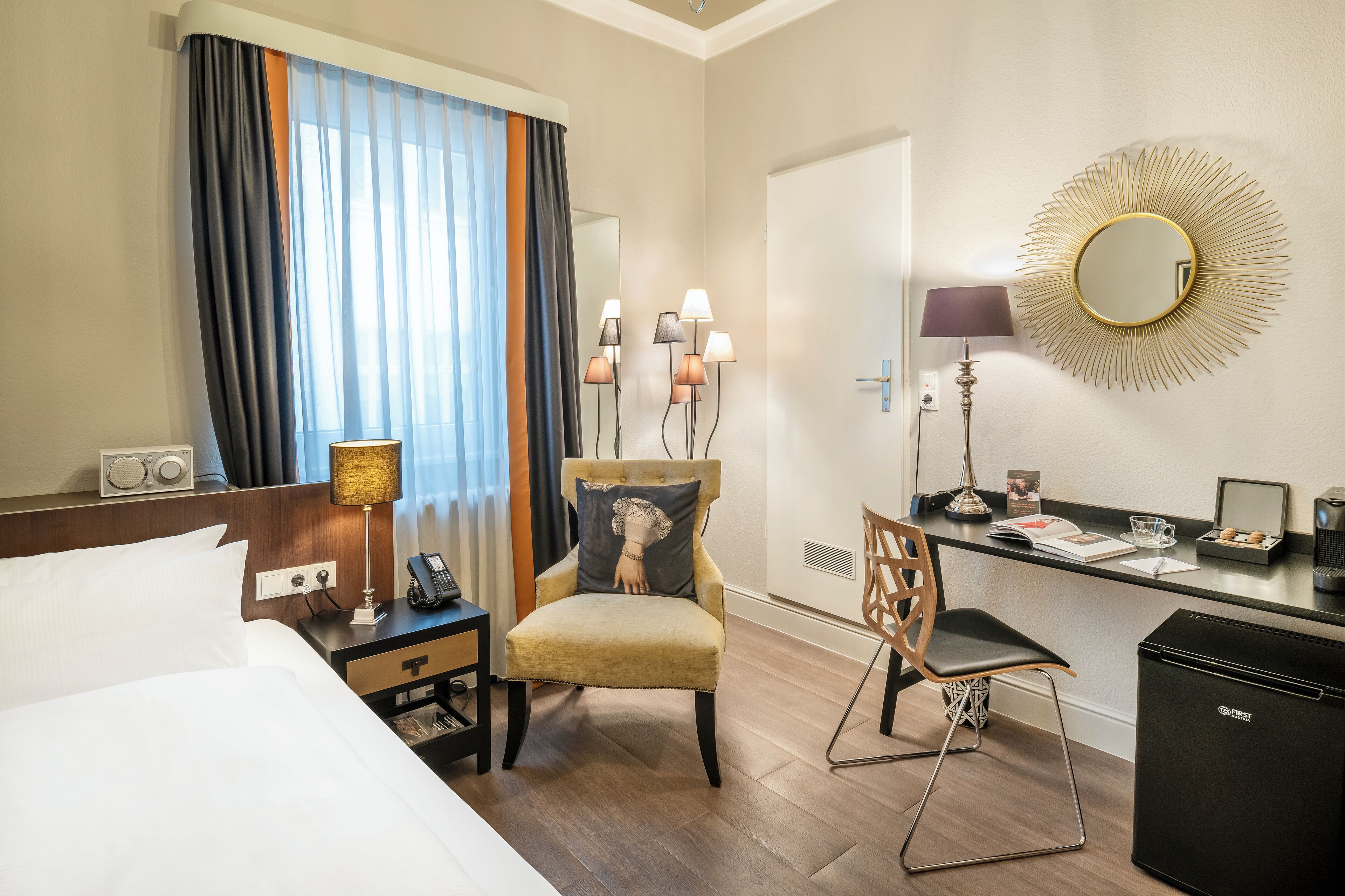 Ein Hotelzimmer mit einem Bett, einem Schreibtisch mit Stuhl, einem kleinen Kühlschrank, einer Lampe, einem Spiegel und einem Fenster mit Vorhängen.