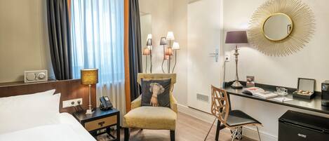 A hotel room with a bed, a desk with a chair, a small refrigerator, a lamp, a mirror, and a window with curtains.