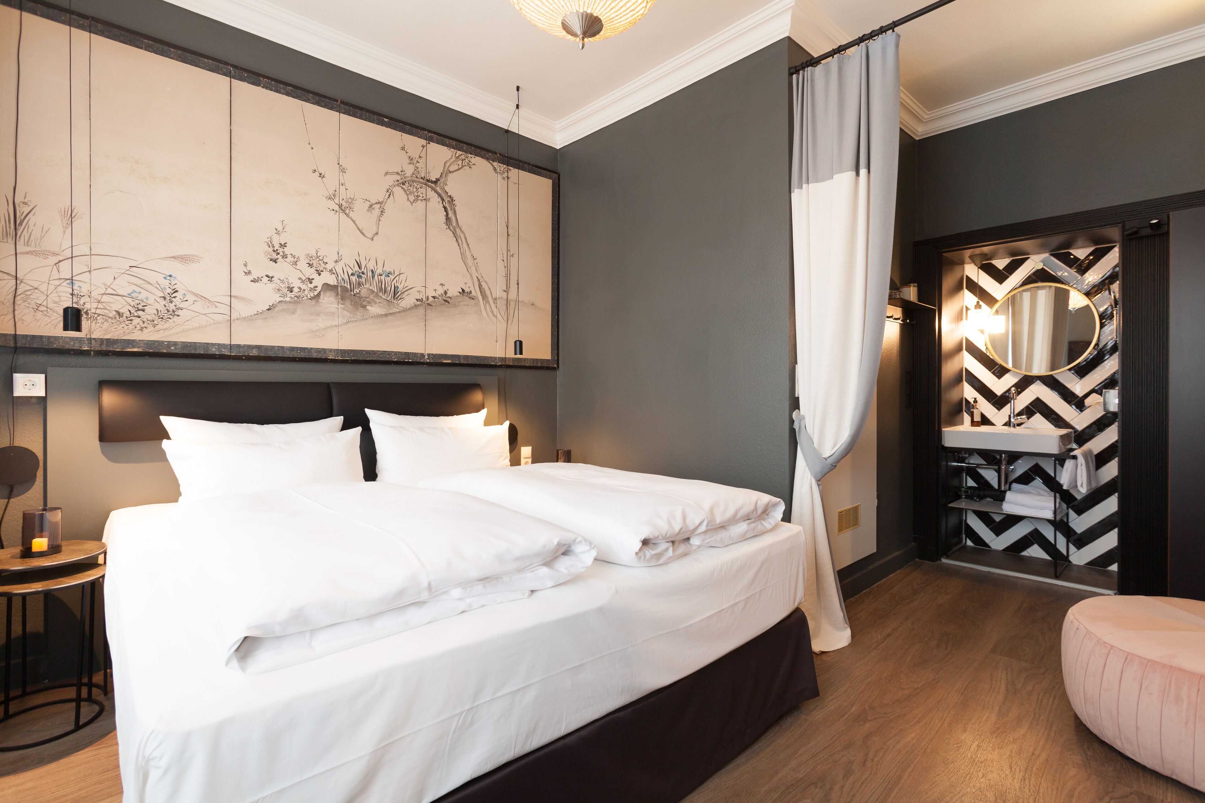 A modern hotel room with a large bed, a bedside table with a lamp, a wall-mounted artwork, and a bathroom with a mirror and towel rack.
