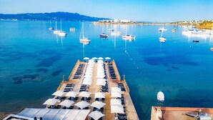 Un muelle con filas de tumbonas y sombrillas, barcos amarrados en el agua, y una costa lejana con edificios.