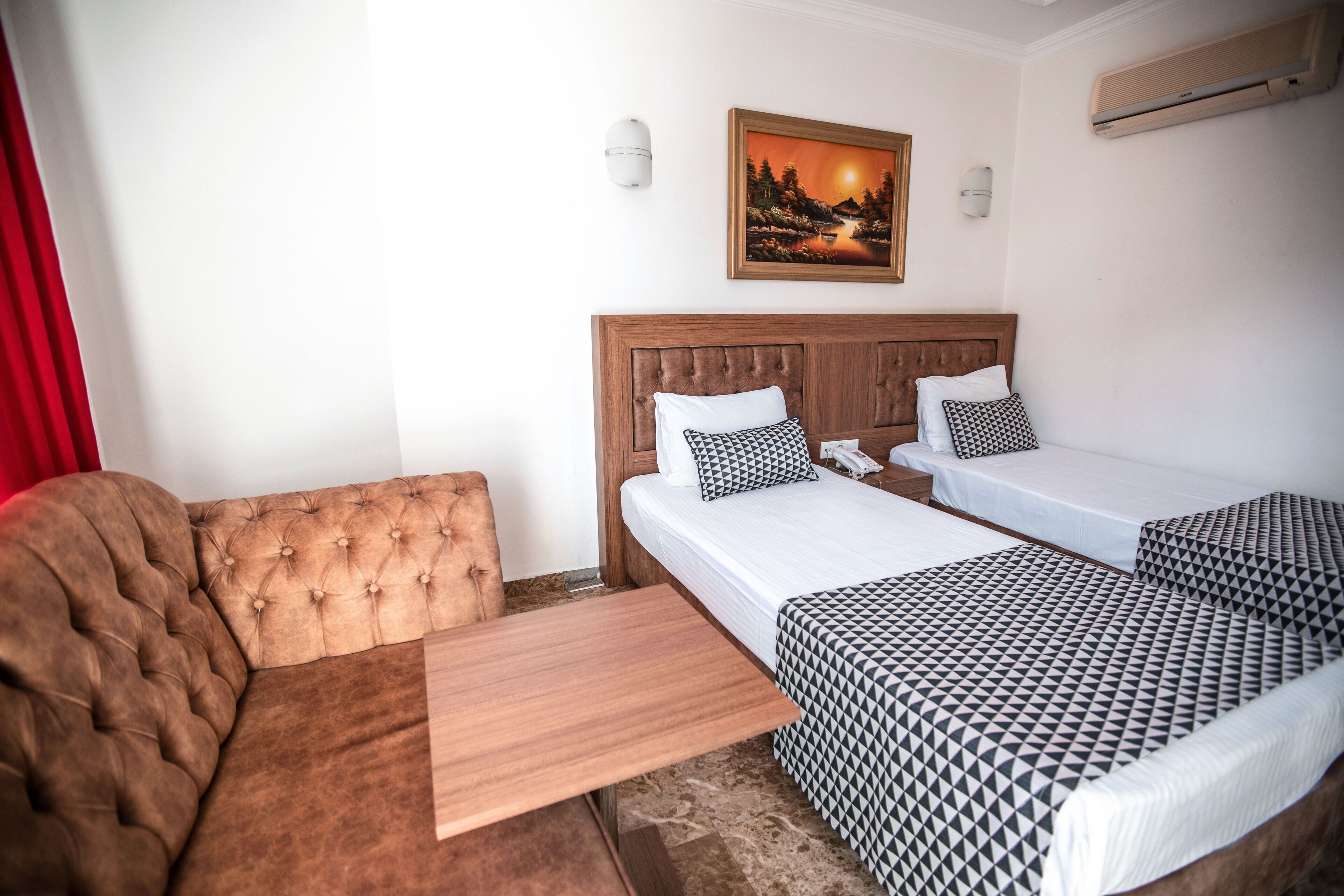 Une chambre d’hôtel avec une tête de lit en bois, deux lits avec une literie à motifs, un banc en bois, un rideau rouge et un climatiseur fixé au mur.