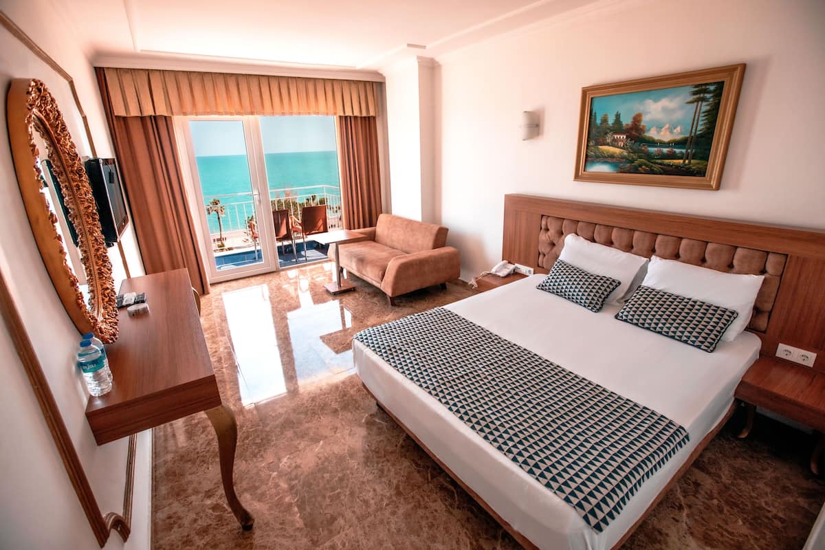 A hotel room with a large bed, a sitting area with a view of the ocean, a painting on the wall, and a wooden desk.