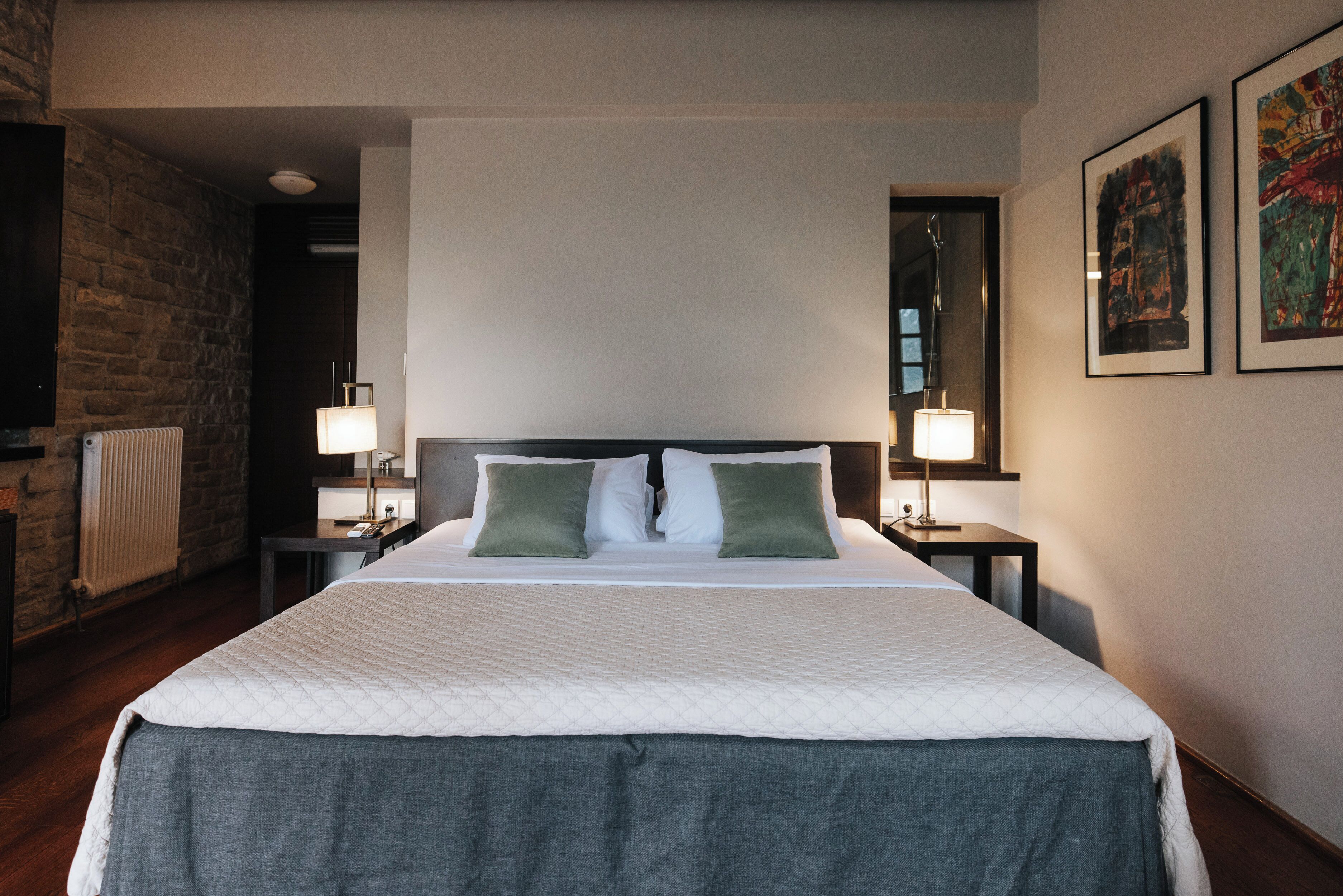 A neatly made bed with a quilted white cover and two green pillows, flanked by two bedside tables with lamps. A brick wall and a radiator are visible to the left.