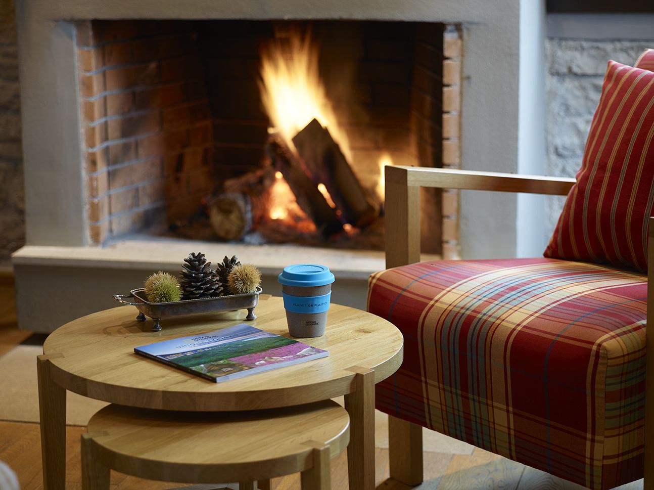 A cozy room with a fireplace, a round wooden table with a book and a coffee cup, and a plaid sofa.