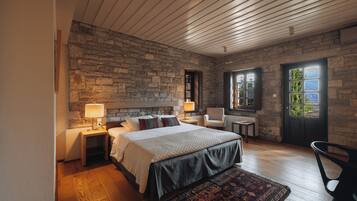 Un dormitorio con pared de piedra, techo de madera, una cama con colcha oscura, una mesita de noche con lámpara y una silla.