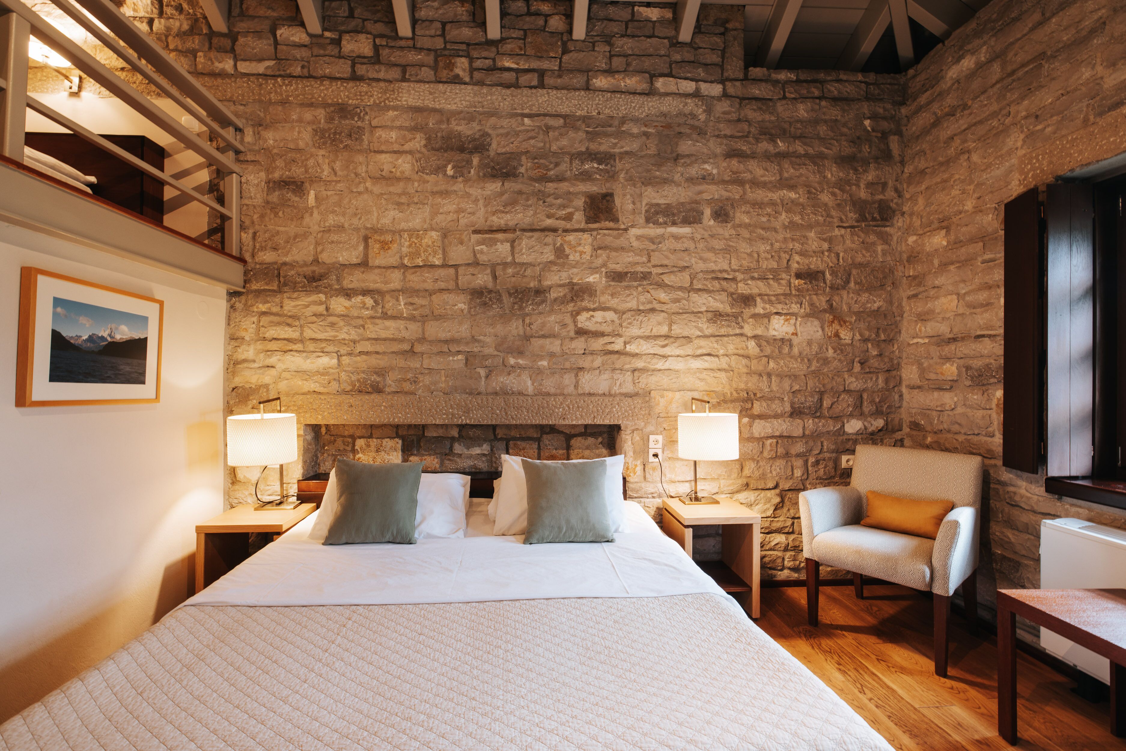 Un dormitorio con pared de piedra, una cama con almohadas, dos mesitas de noche con lámparas, una silla y una pequeña mesa.