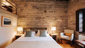Un dormitorio con pared de piedra, una cama con almohadas, dos mesitas de noche con lámparas, una silla y una pequeña mesa.