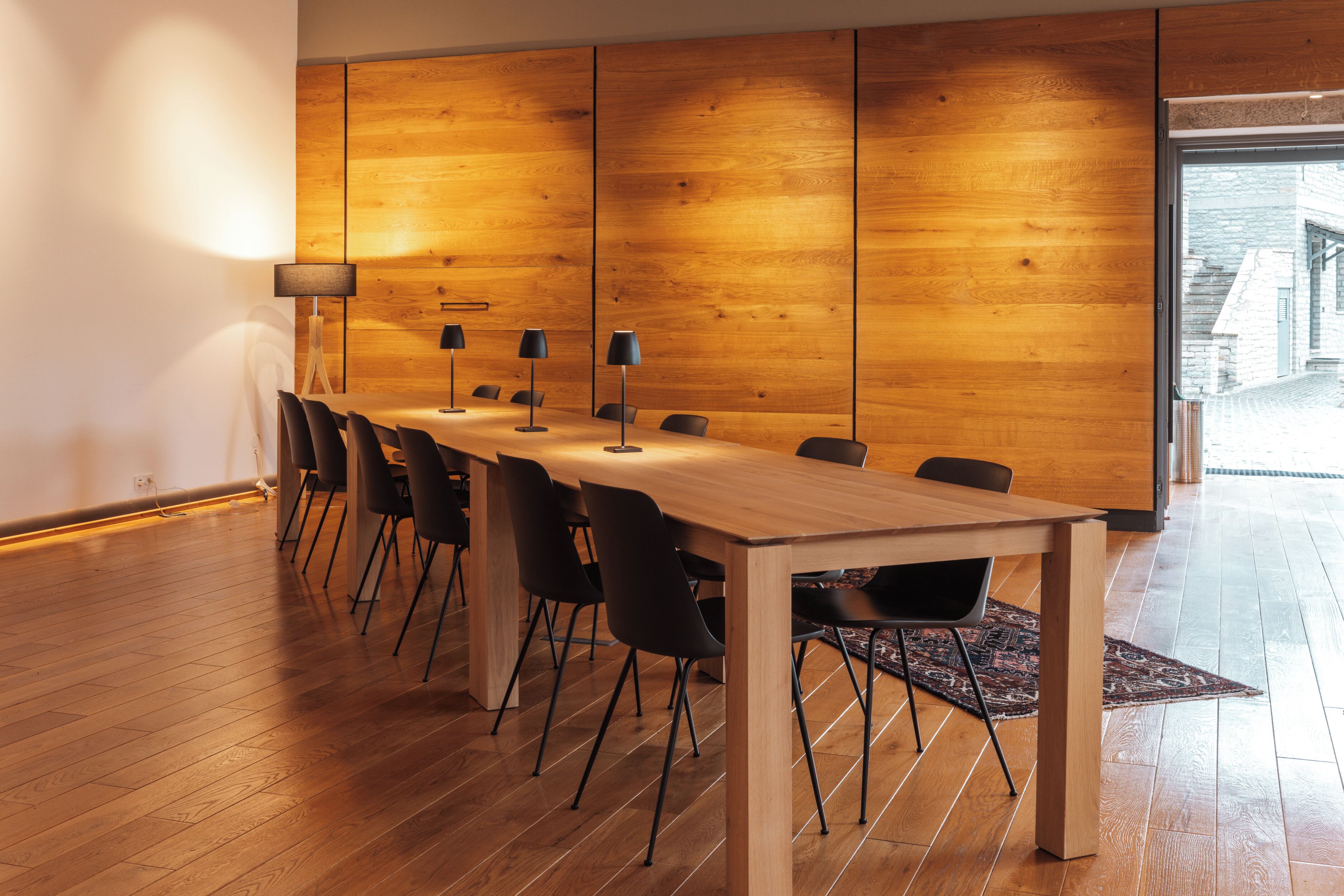 A conference room with a wooden table and chairs, a patterned rug, and a large window offering a view of the outside.