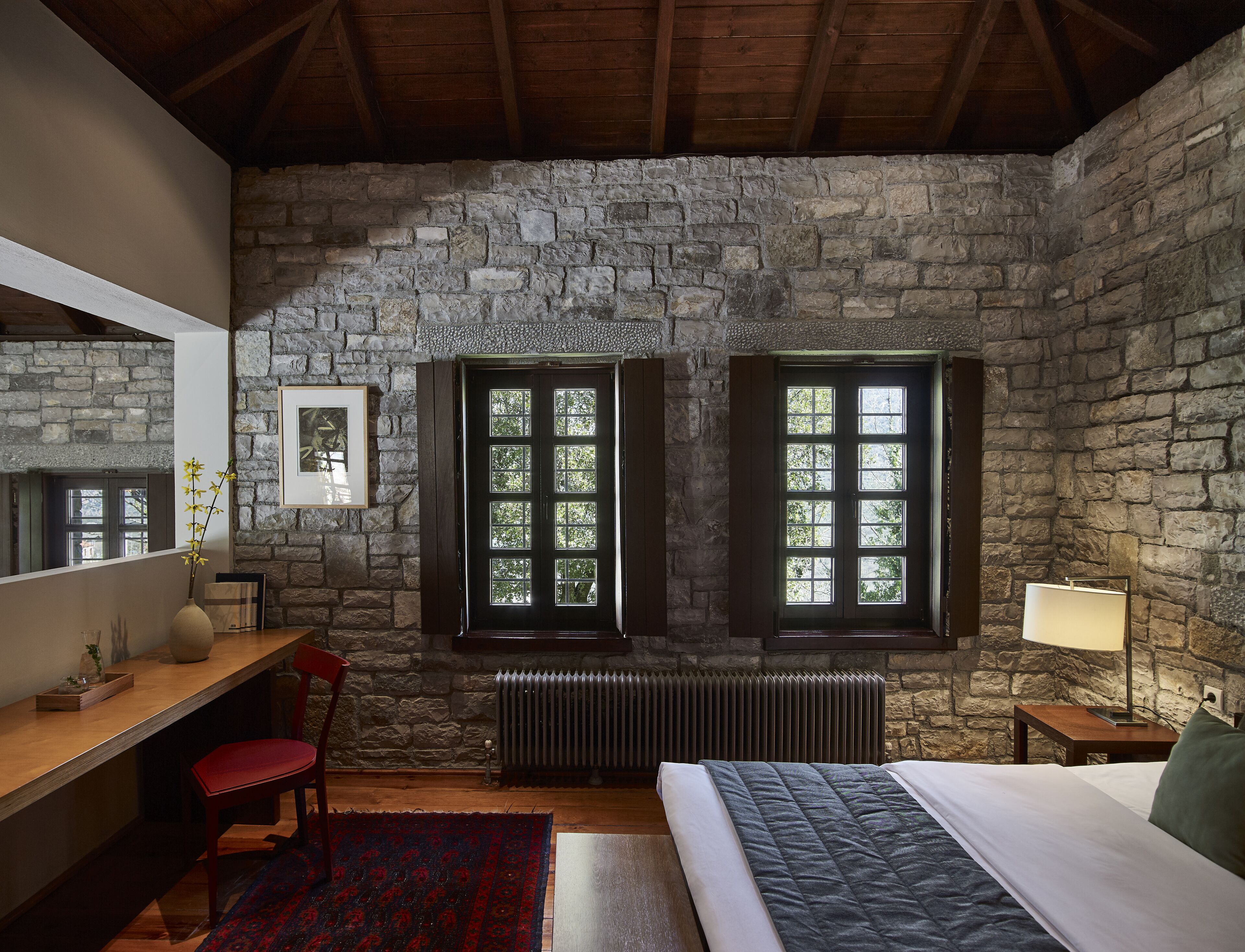 A bedroom with stone walls, a wooden ceiling, a bed, a desk, a chair, a radiator, and two windows with shutters.
