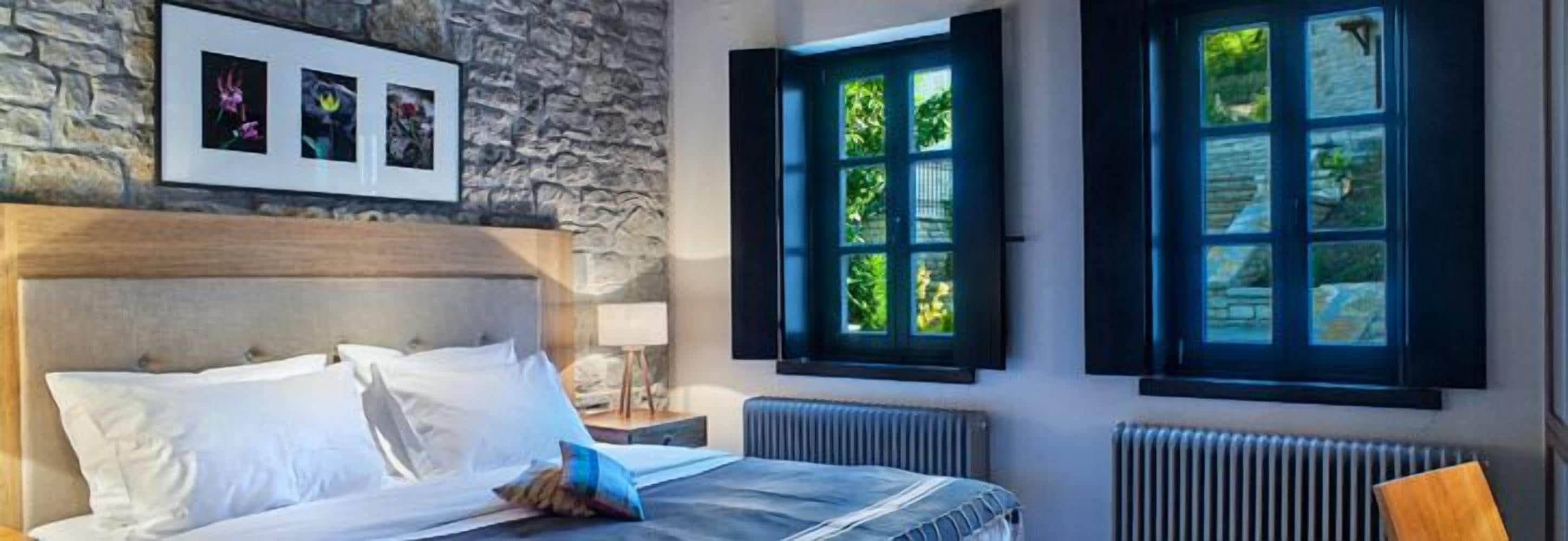 Habitación de hotel con cama, pared de piedra, ventana con contraventanas y radiador.