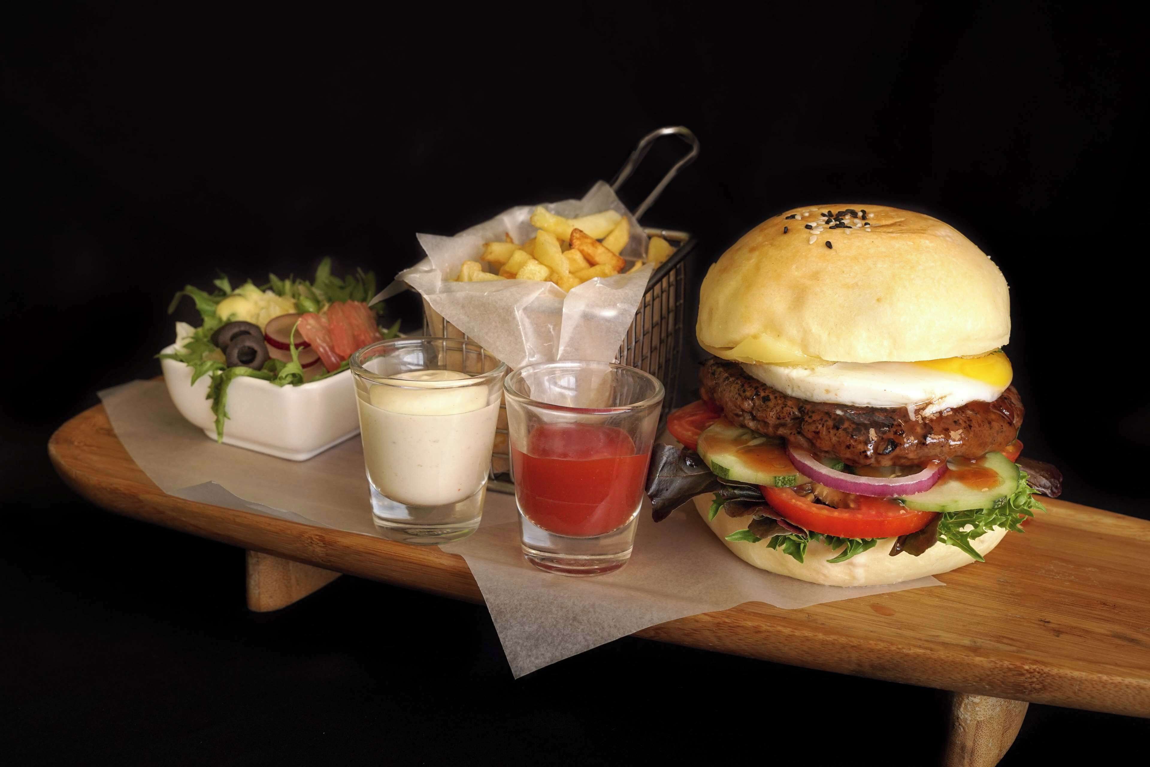 Un plato con una hamburguesa, papas fritas, ensalada y dos salsas sobre una bandeja de madera.