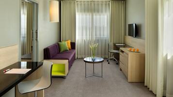 Habitación de hotel con sofá, escritorio, silla, mesita con un florero, televisión y ventana con cortinas.