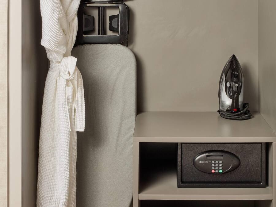 A neatly organized hotel room with a built-in ironing station, a robe hanging on a rack, a pillow, a small bag, and slippers.