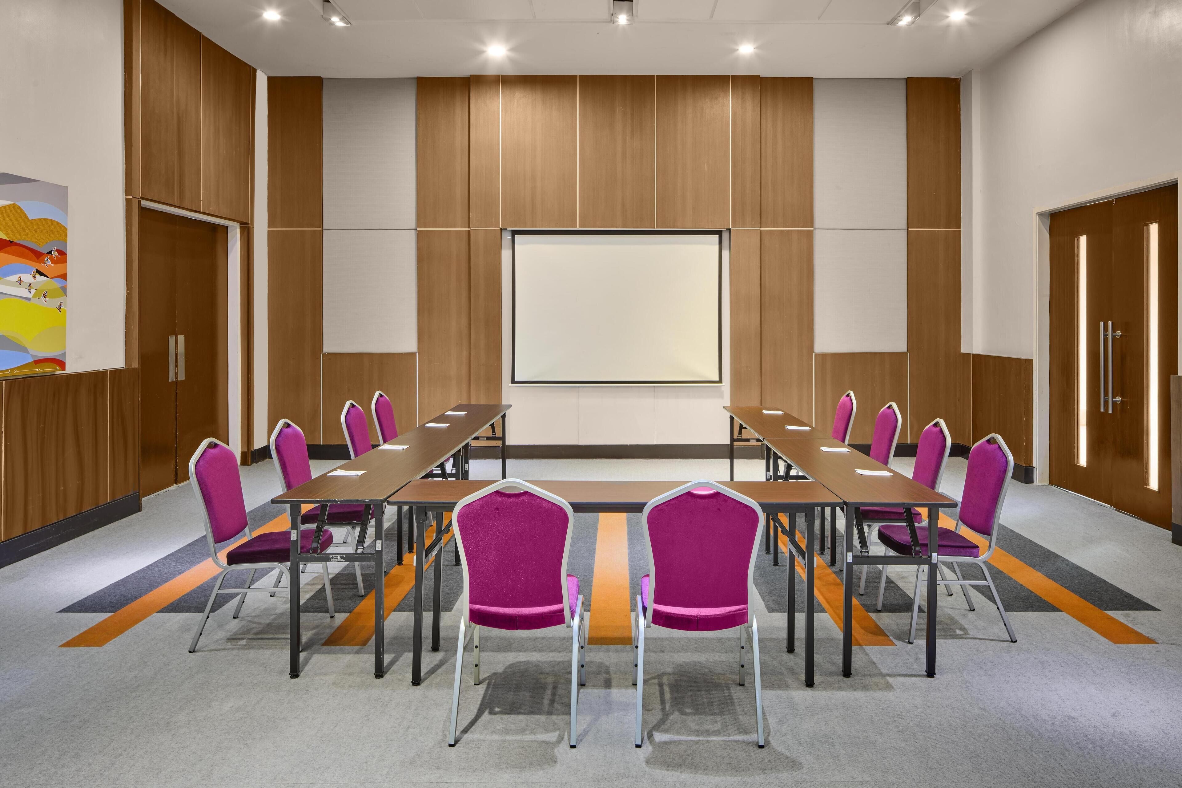 A conference room with a long table, purple chairs, and a large screen.