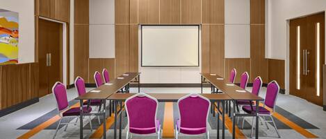 Un salón de conferencias con una larga mesa, sillas moradas y una pantalla grande.