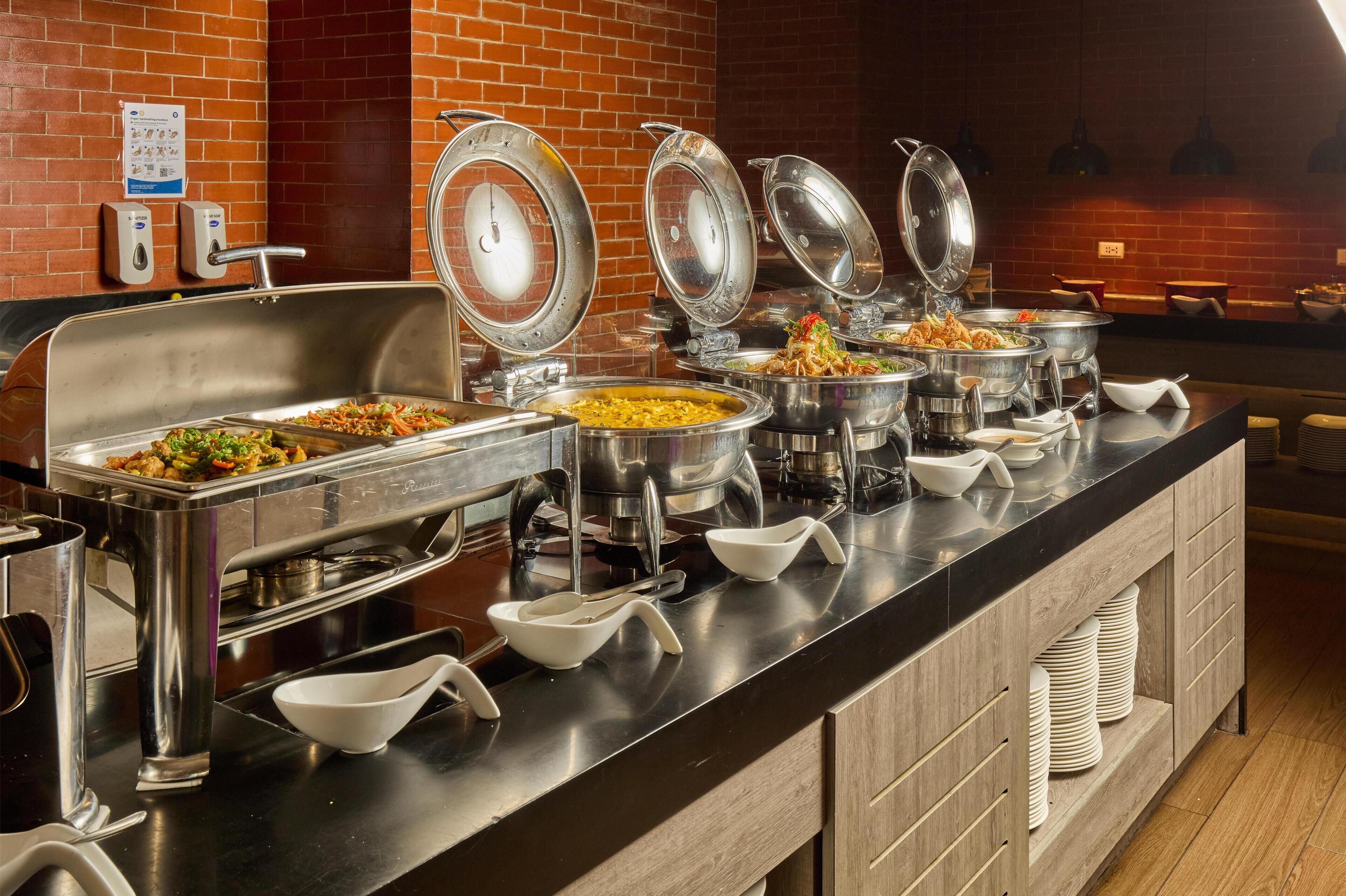 Un buffet avec divers plats présentés dans des récipients en métal, un distributeur de savon liquide et un thermomètre fixé au mur.