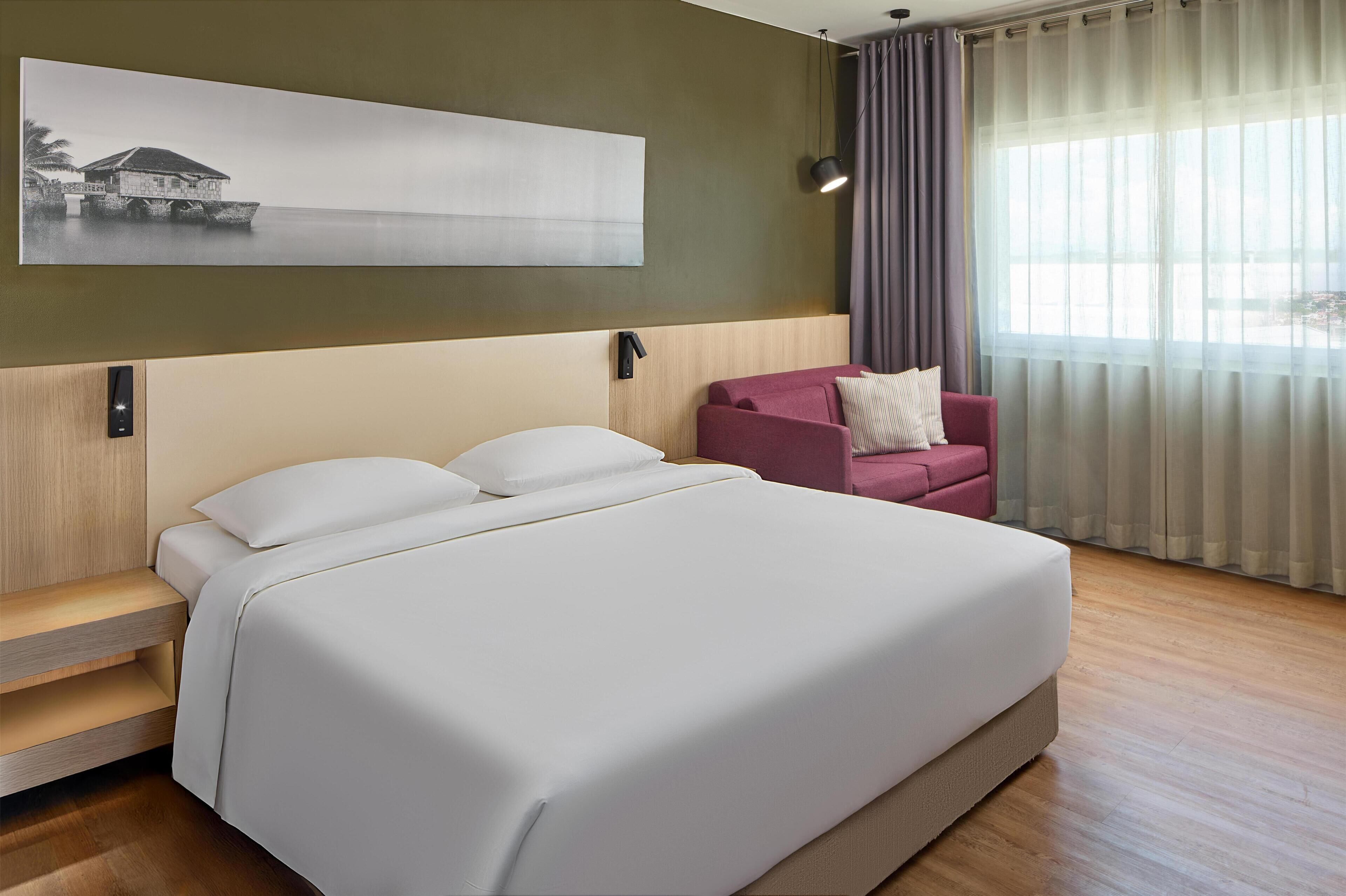 Una habitación de hotel con cama, una silla, un cuadro en la pared y una ventana con cortinas.