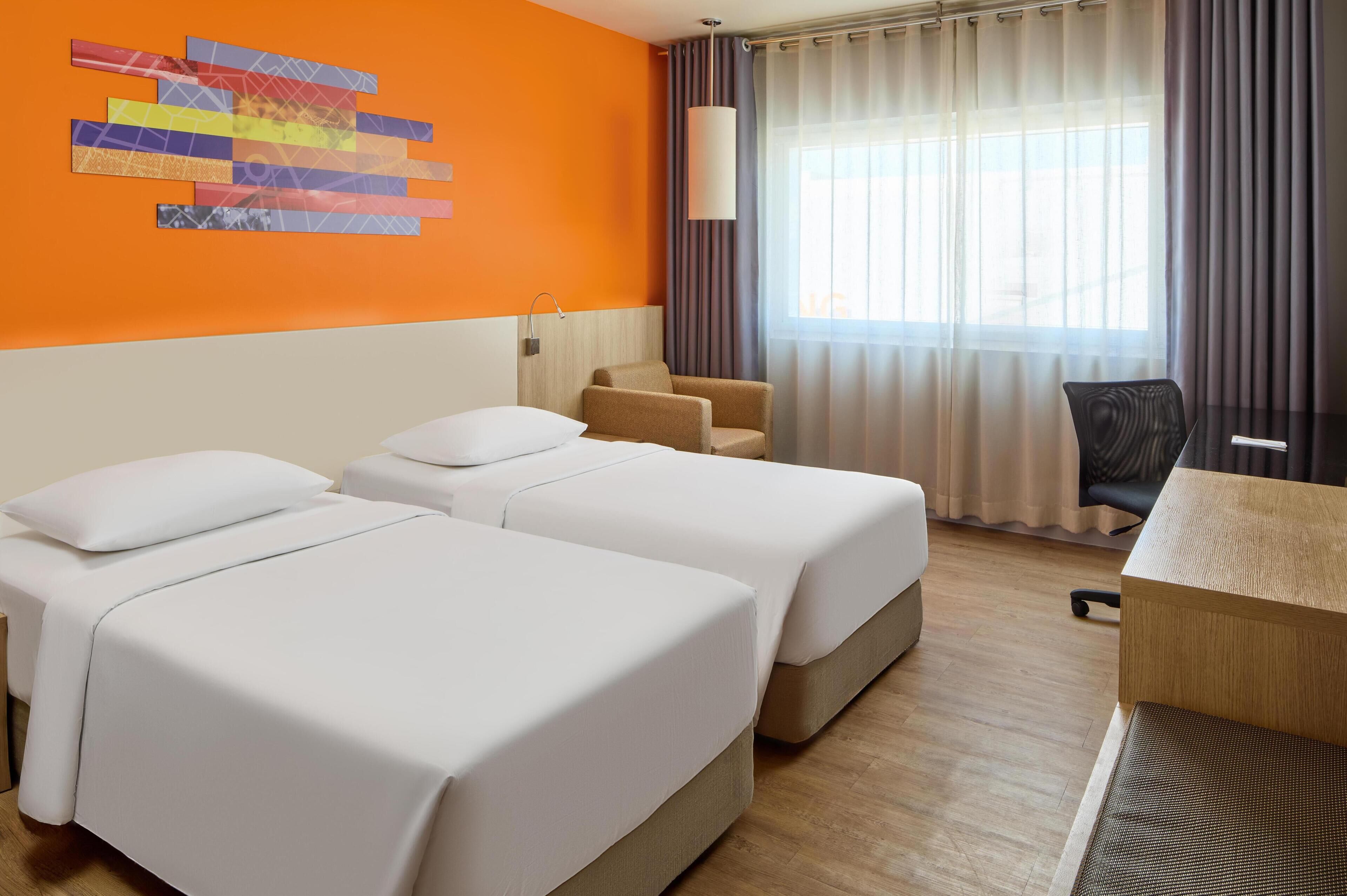 A hotel room with two beds, a desk, a chair, and a large window with curtains.