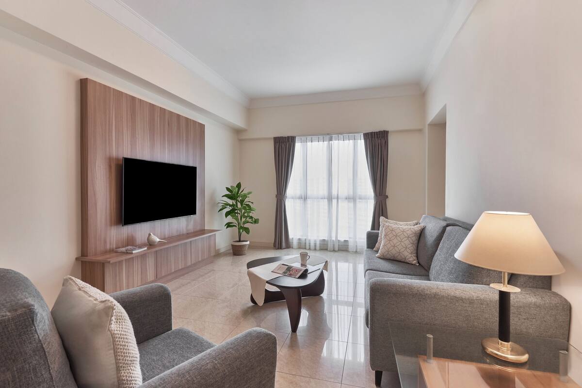 A modern living room with a flat-screen TV, grey sofas, a coffee table, a lamp, and a potted plant.