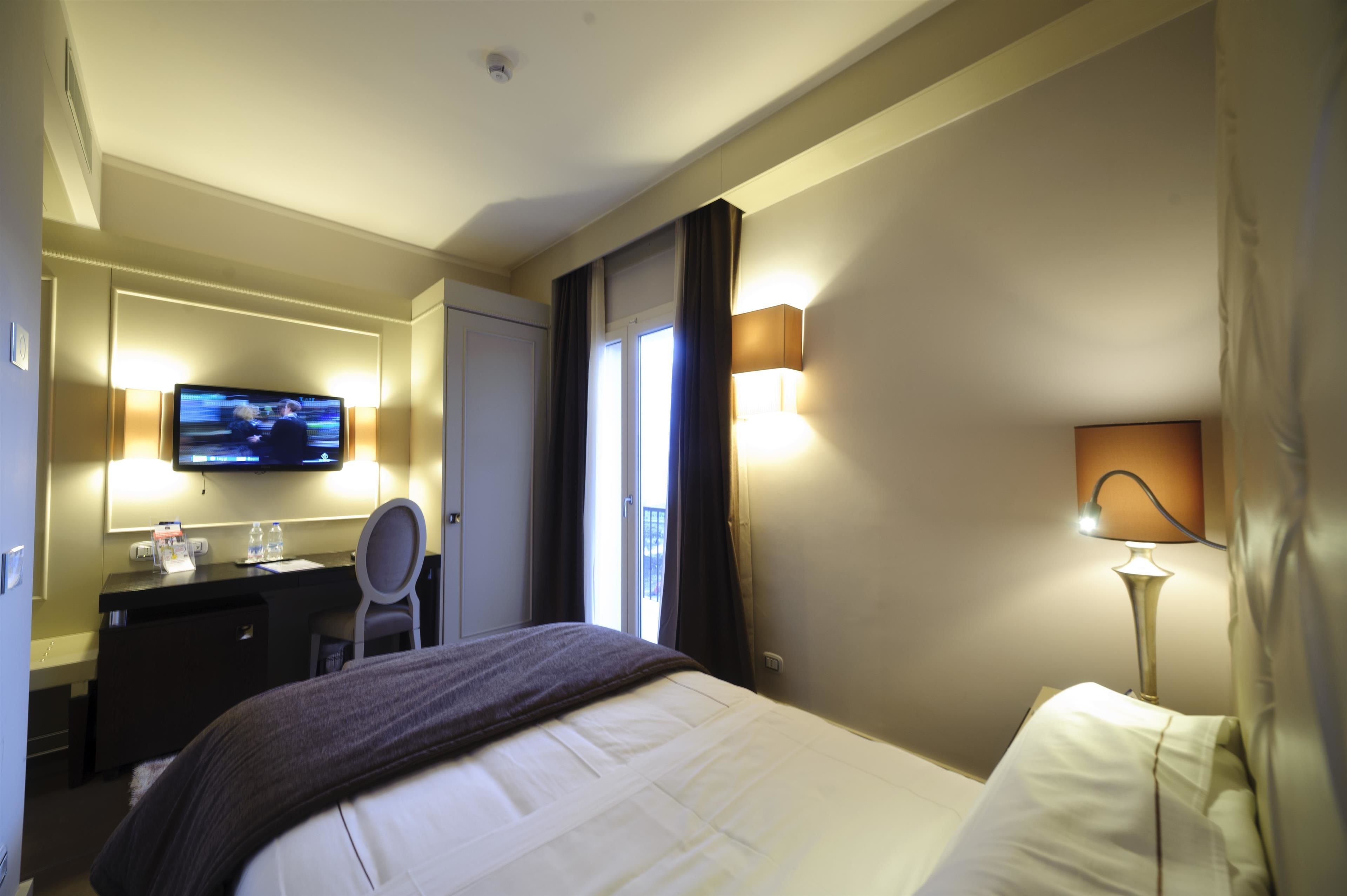A hotel room with a bed, a desk with a television, a chair, and a lamp.