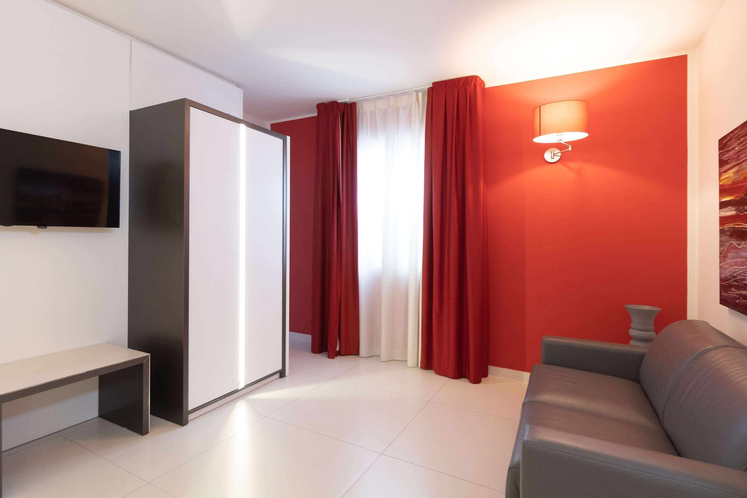 Una sala de estar moderna con un sofá gris, un televisor de pantalla plana montado en una pared blanca, un armario con puerta corrediza y una pared de acento roja con una lámpara de pared.