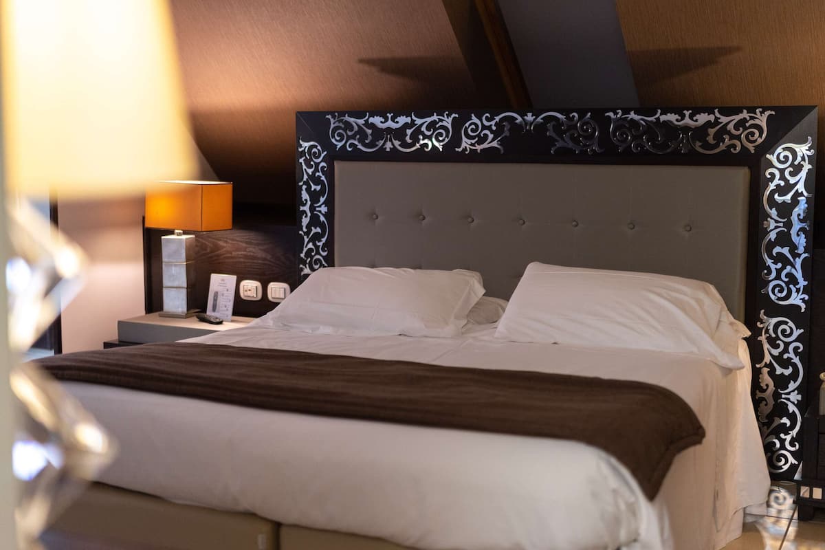A neatly made bed with white linens and a brown throw blanket, a bedside table with a lamp, and a decorative headboard with intricate patterns.