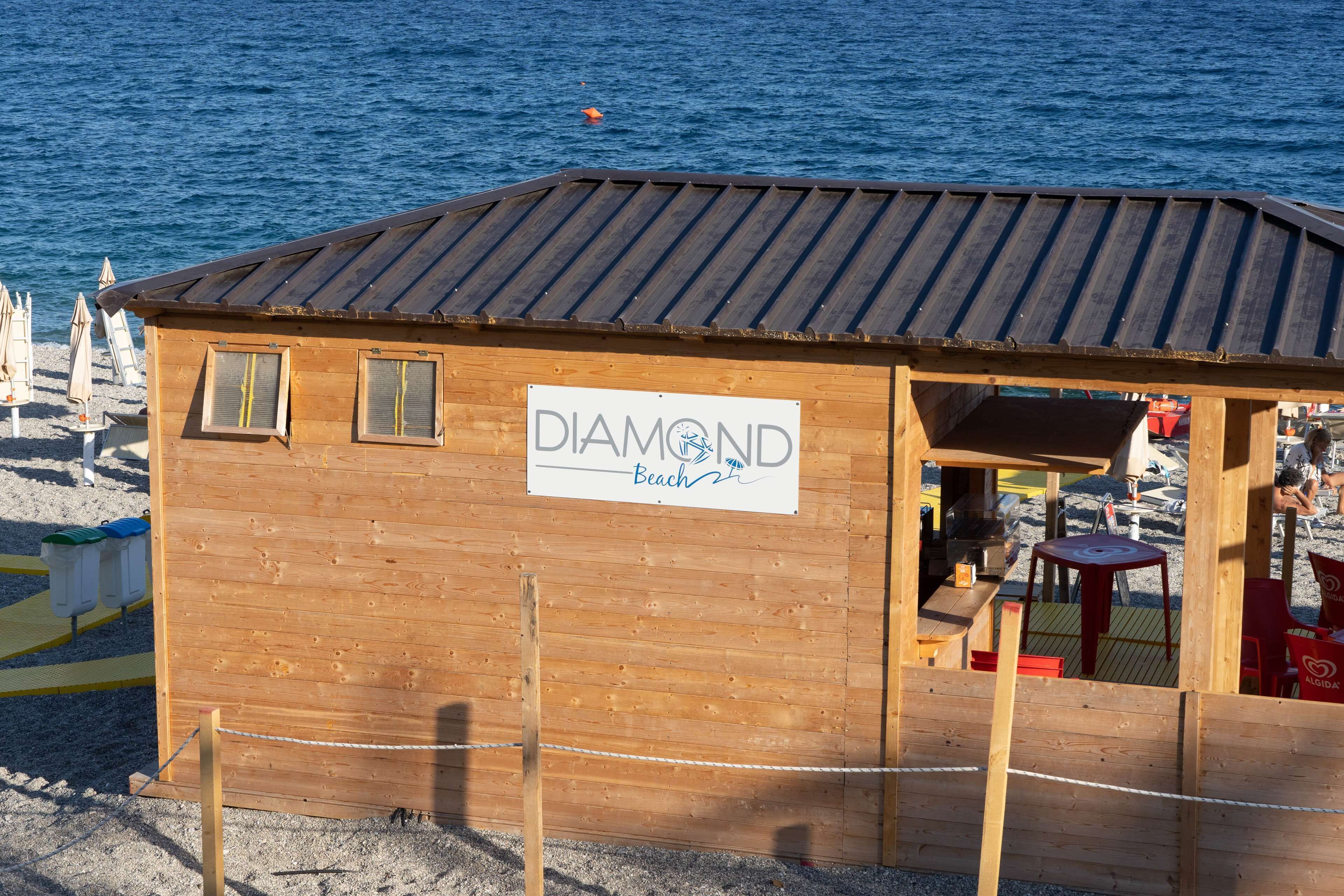 A wooden beach hut labeled "DIAMOND Beach" with a metal roof, situated on a sandy beach with a view of the sea.