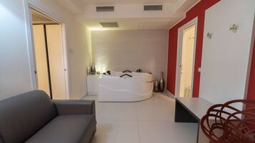 Een moderne badkamer met een bad, toilet en douche. In de aangrenzende woonruimte staat een grijze bank en een rode fauteuil.