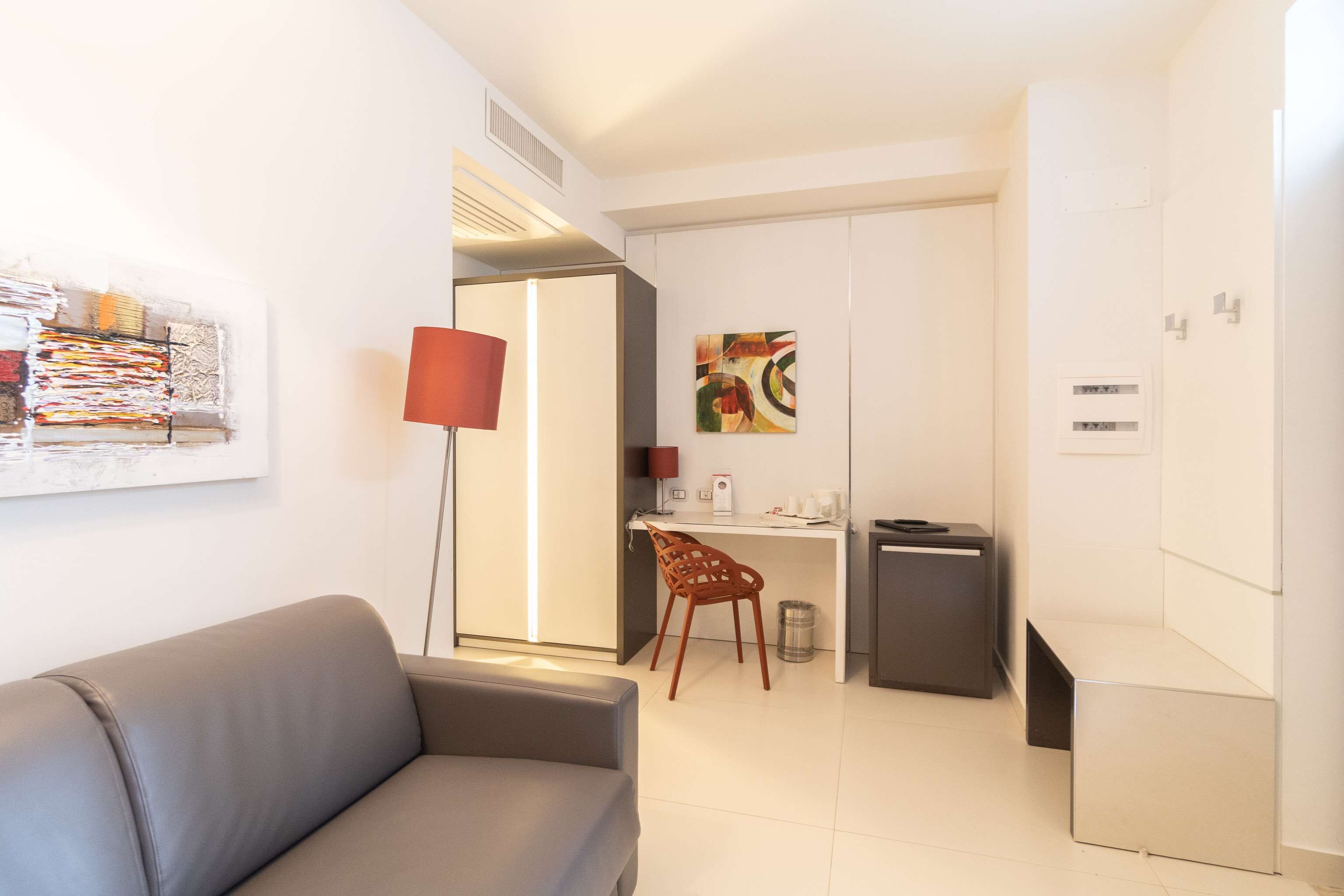 Een moderne hotelkamer met een grijze bank, een bureau met een stoel, een prullenbak en een kleine kitchenette.