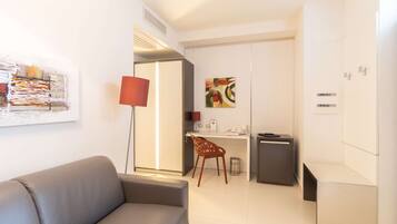 Een moderne hotelkamer met een grijze bank, een bureau met een stoel, een prullenbak en een kleine kitchenette.