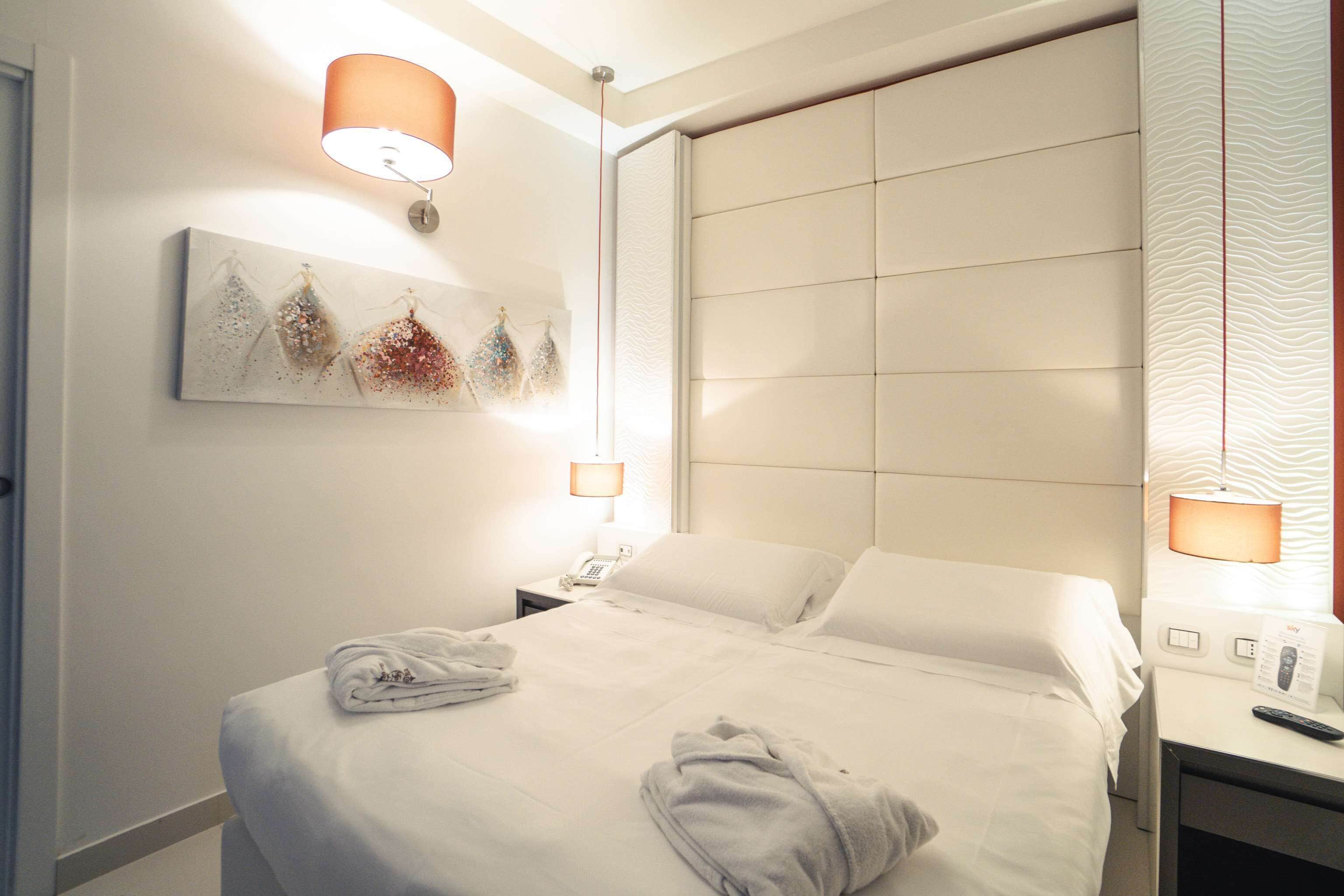A neatly made bed with white linens, a bedside table with a lamp, and a wall-mounted artwork.