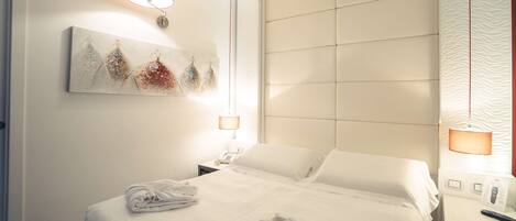 A neatly made bed with white linens, a bedside table with a lamp, and a wall-mounted artwork.