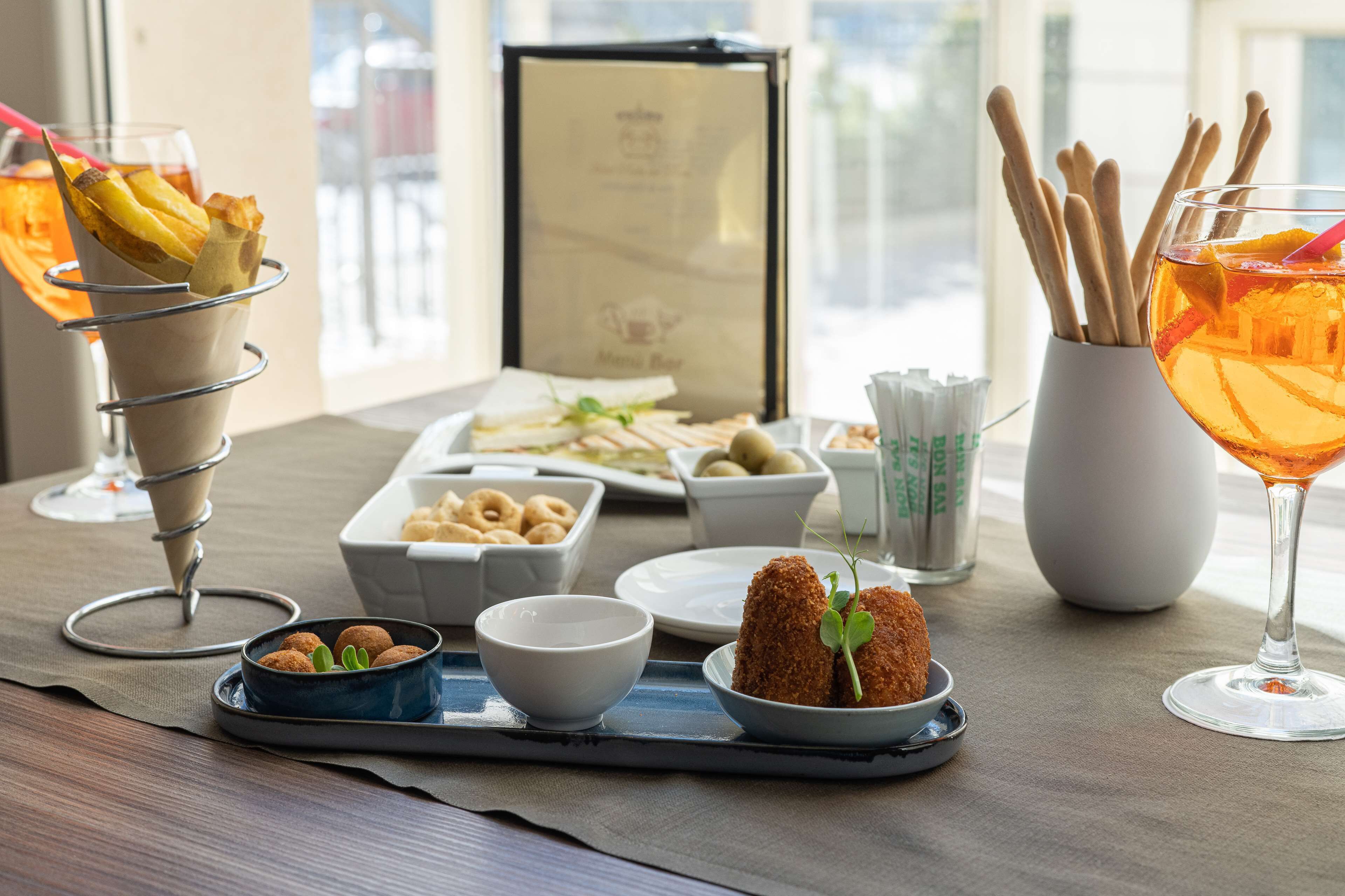 A table with a variety of food items including a dessert in a metal cup, a plate with bread rolls, a bowl of nuts, and a glass of orange drink.