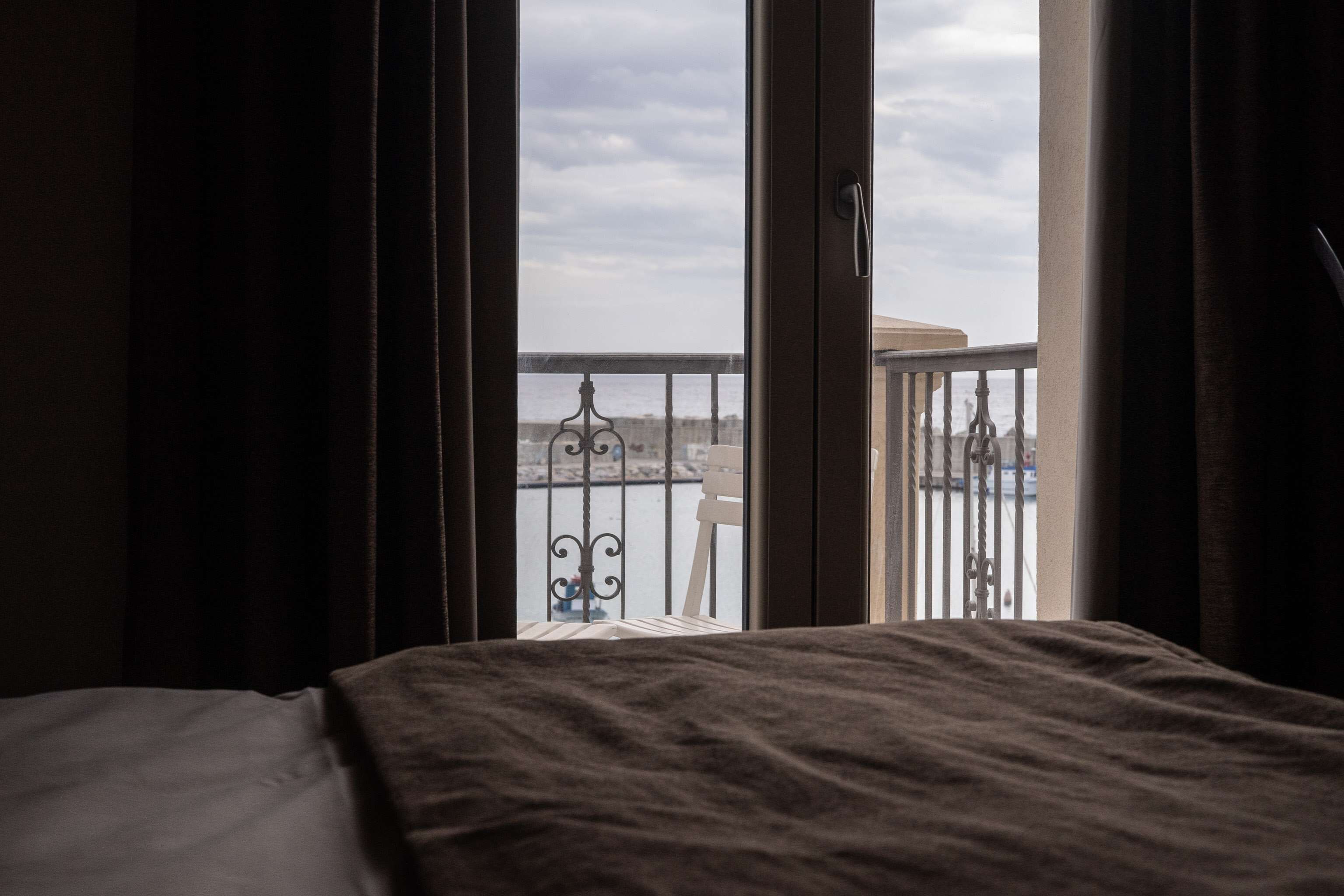 A hotel room with a bed, a balcony with a railing, and a view of the sea and a cityscape.