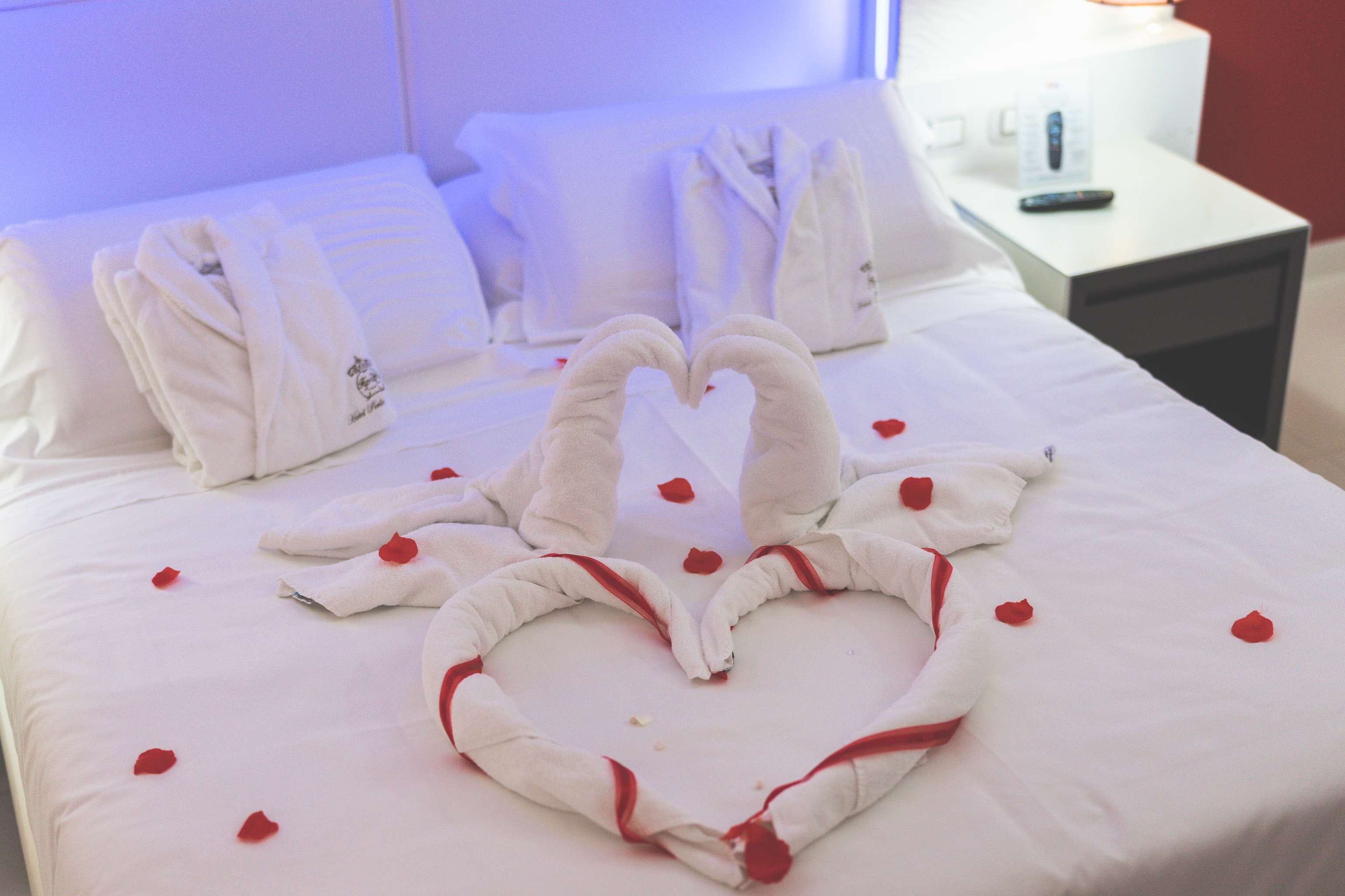 Een net opgemaakt bed met handdoeken gerangschikt in de vorm van een hart en rozenblaadjes verspreid over het bed.