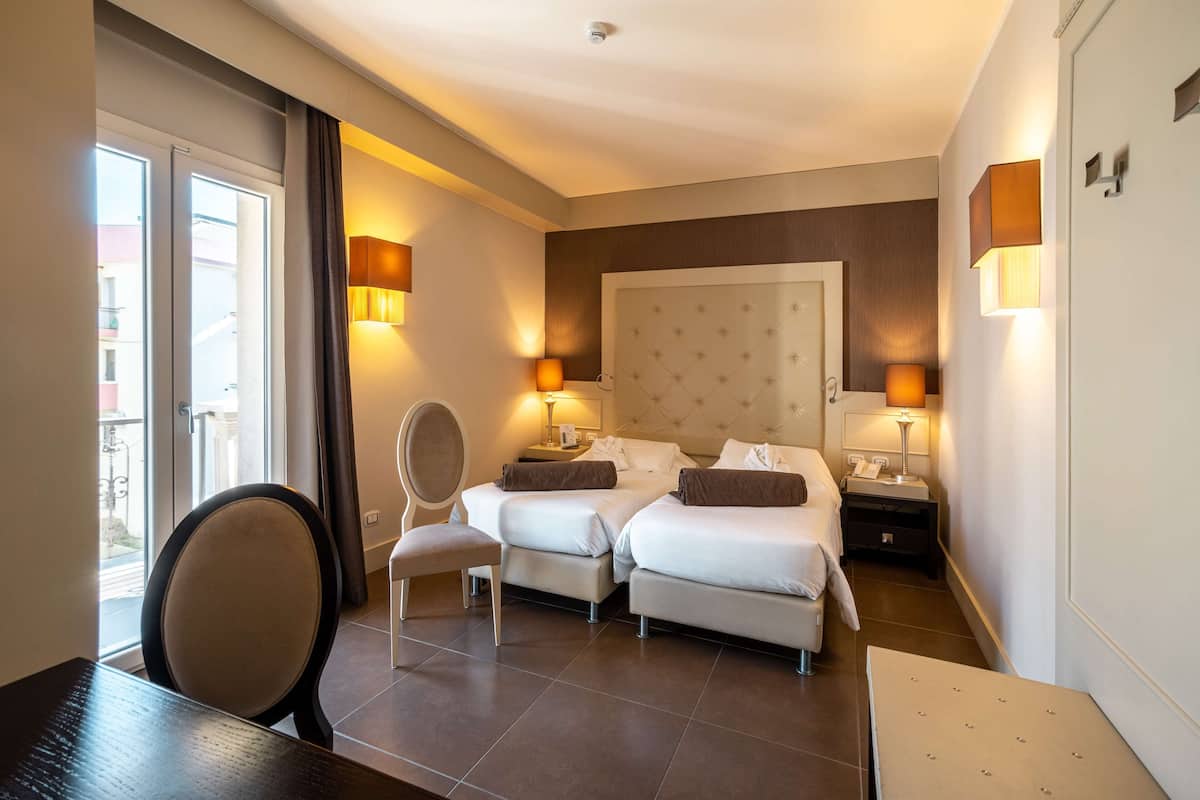 A hotel room with two beds, a chair, a desk, and a balcony.