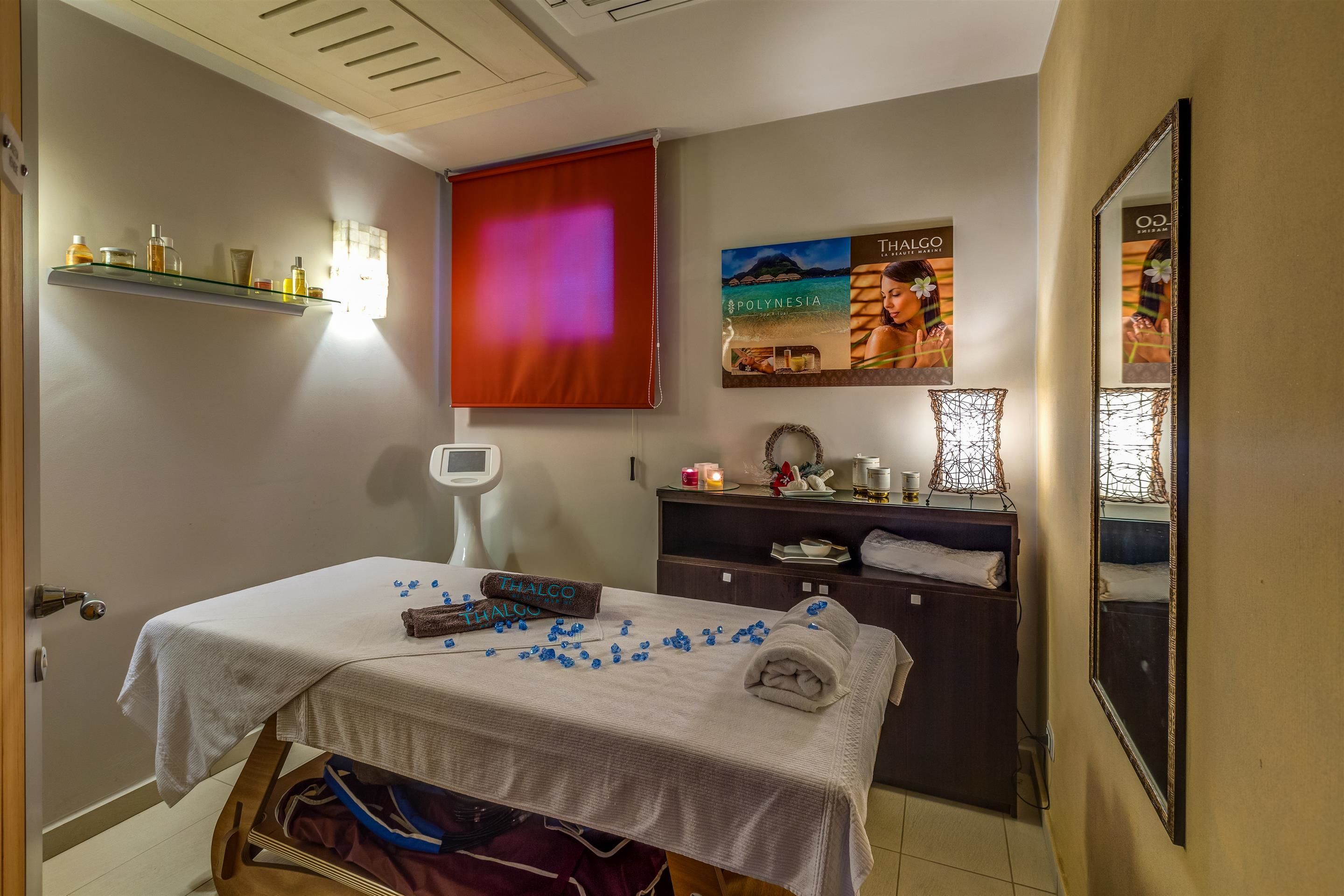 Quarto com mesa de massagem, prateleira com artigos de higiene, balança digital, pôster e espelho.