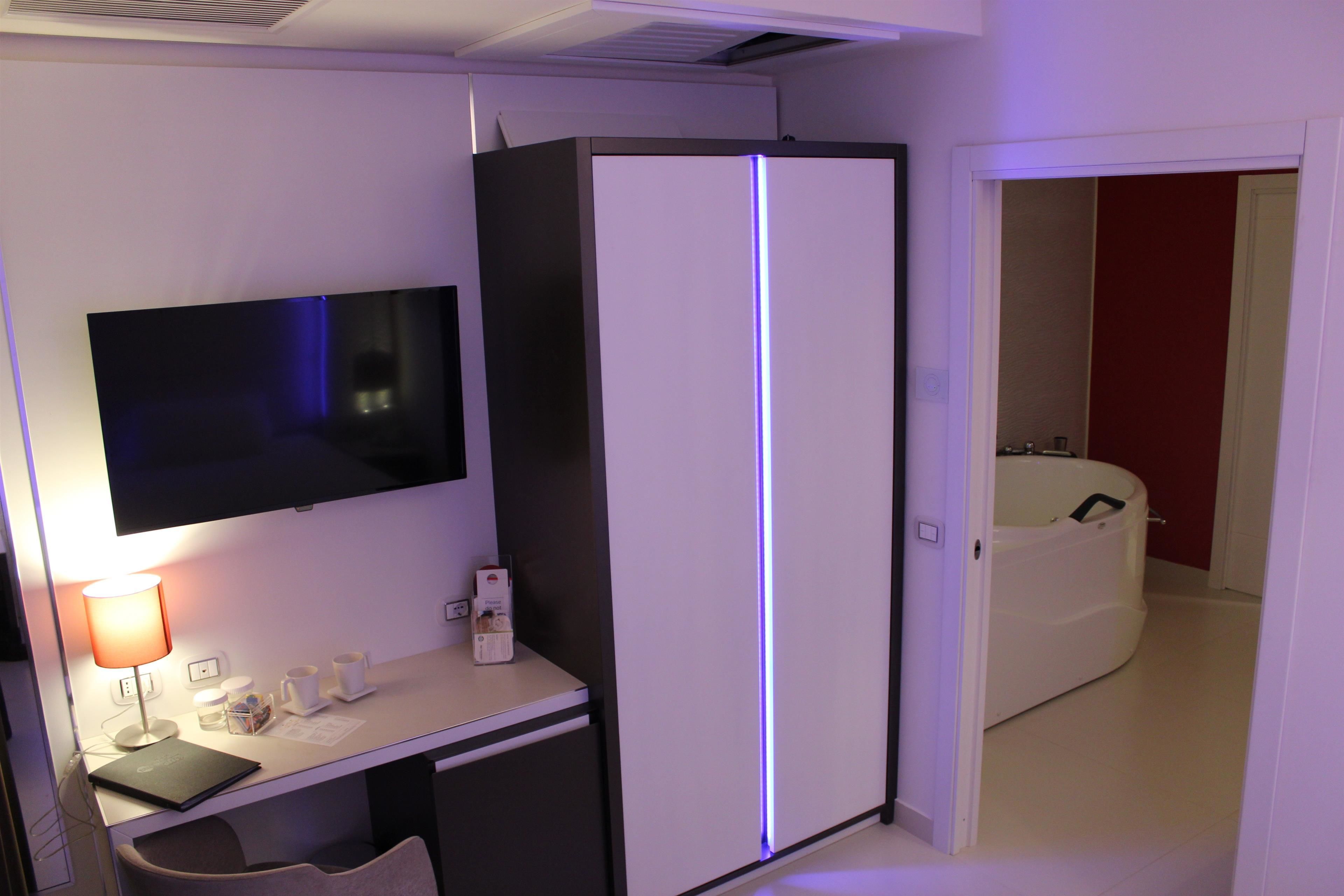 Quarto de hotel com televisão de tela plana, escrivaninha com luminária, geladeira e guarda-roupa branco.