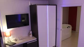 Quarto de hotel com televisão de tela plana, escrivaninha com luminária, geladeira e guarda-roupa branco.