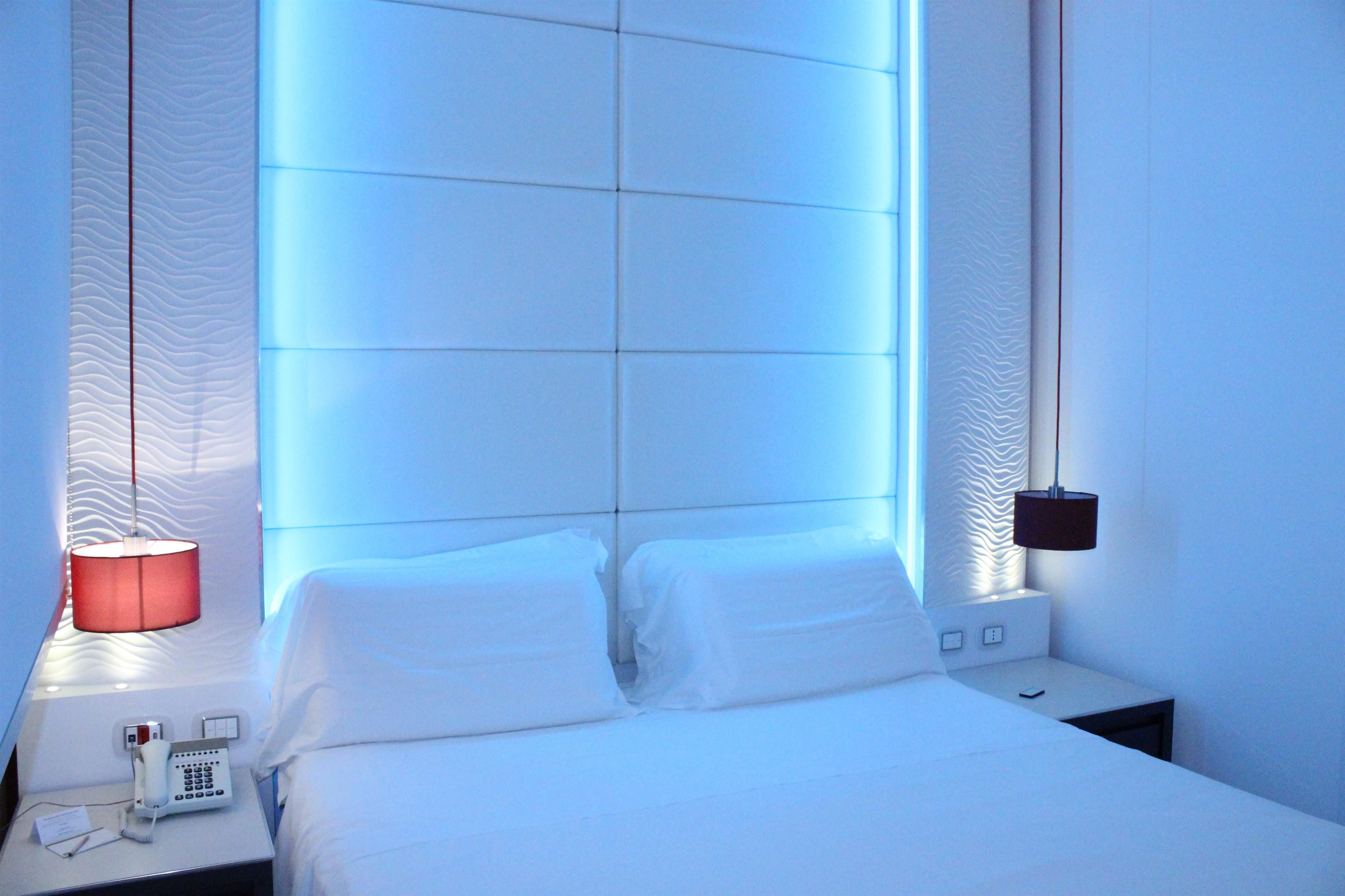 A hotel room with a bed, a bedside table with a lamp, a phone, and a wall-mounted light fixture.